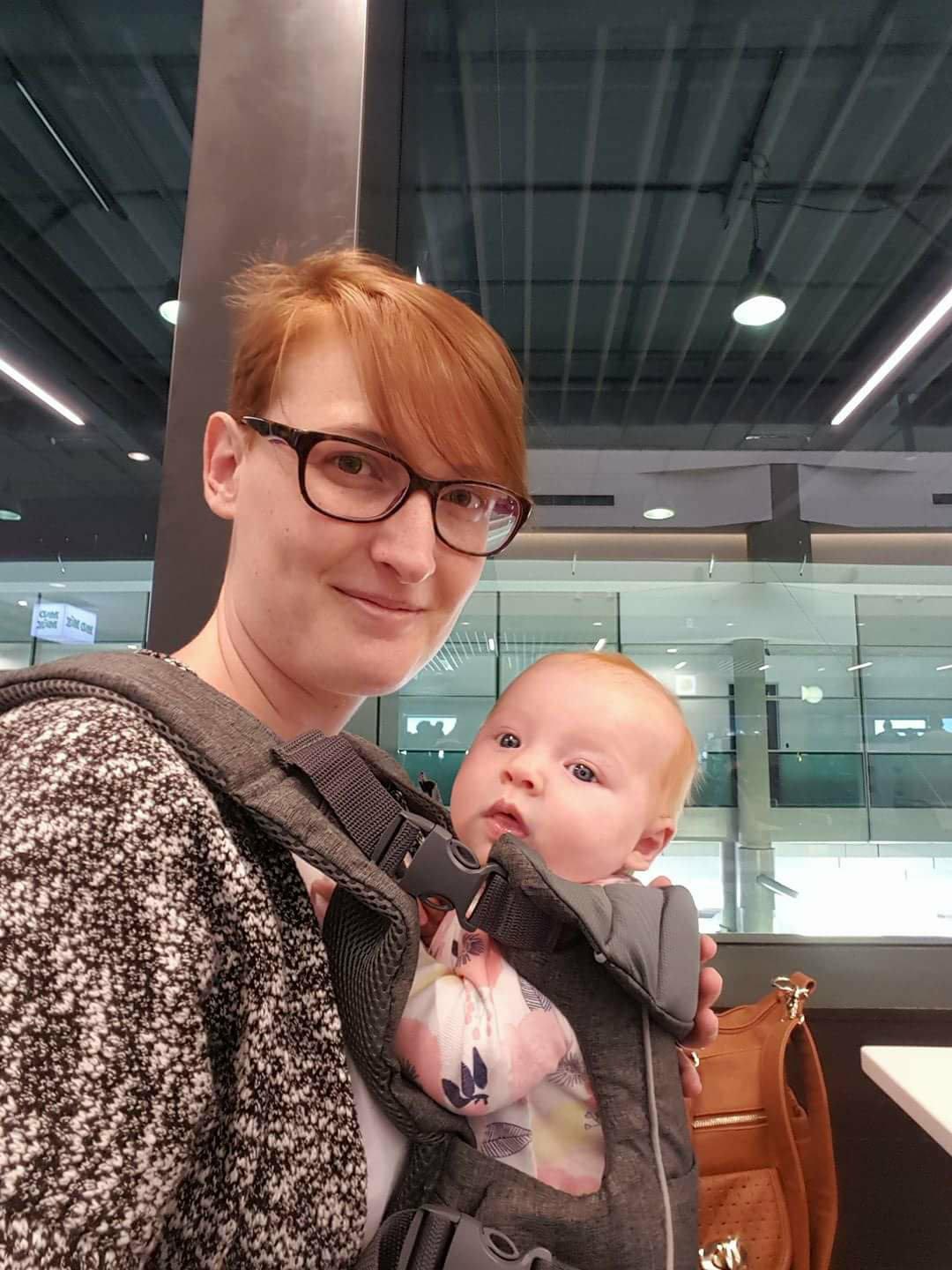 Red haired mother with baby in a carrier smiling at the camera.