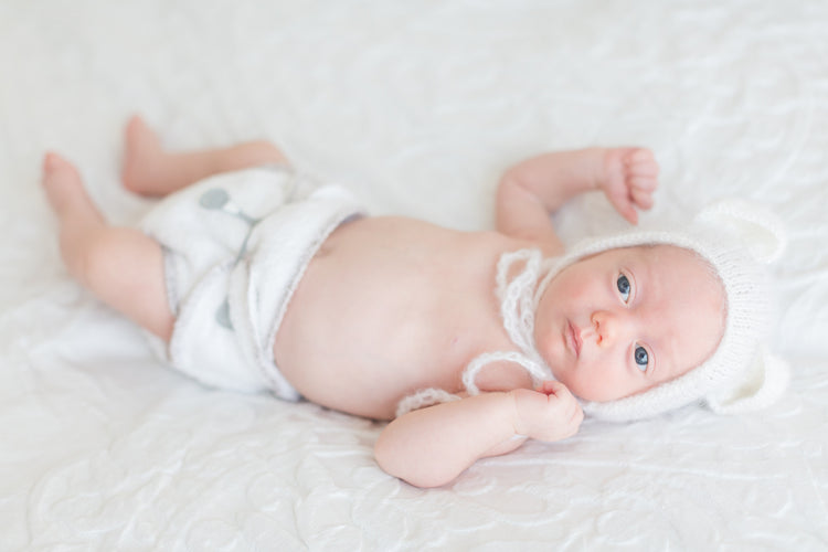 A baby with deep blue eyes waring a Bubblebubs Bambam cloth nappy done up with a grey snappi laying on a soft bed waring a bonbon. The reusable nappy is out of focus, but you can see it fits well.